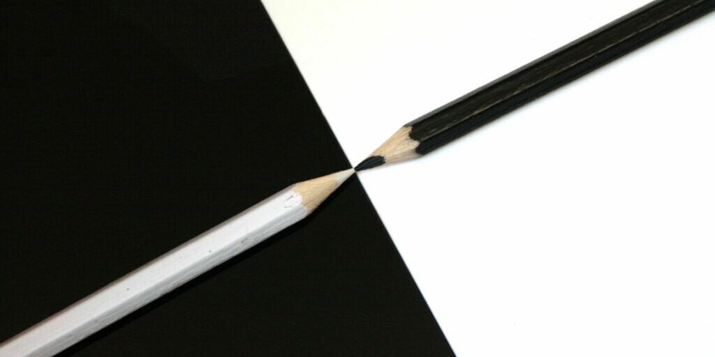 Two pencils are laid down with their tips touching each other. On the left hand side is a white pencil with white lead on a black background, and on the right hand side is a black pencil with black lead on a white background.