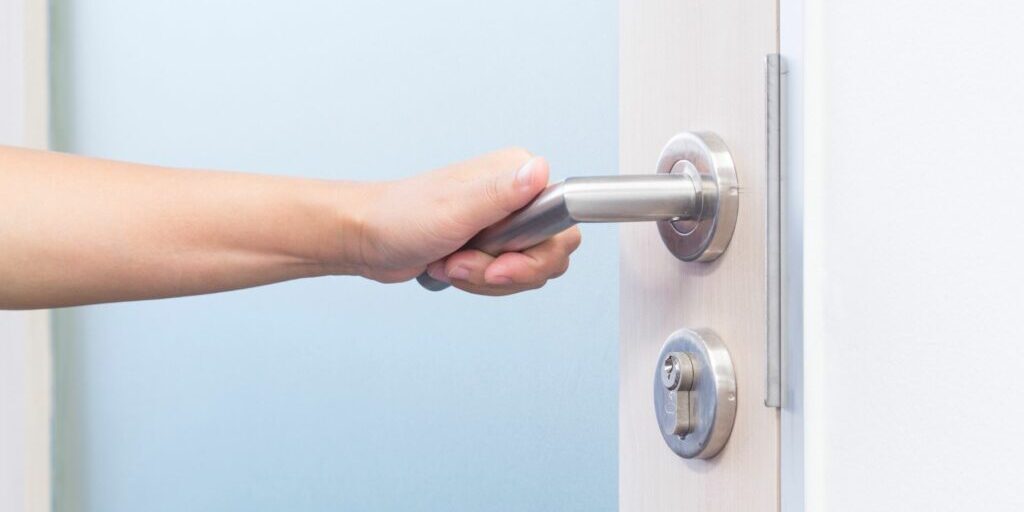 A close up photo of a person's left hand opening up a door by turning a silver door handle.