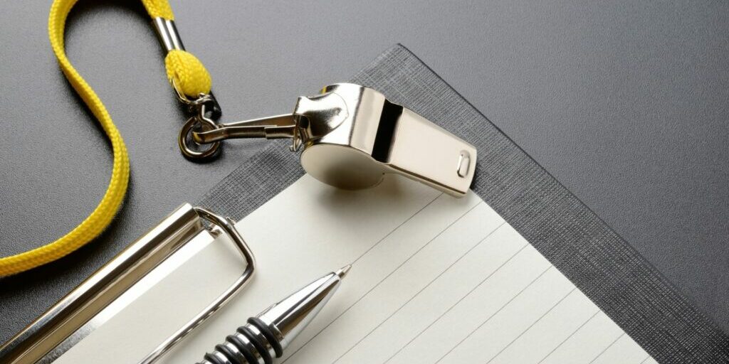 a silver whistle with a yellow strap is laid down on a grey clipboard with white lined paper and a pen on top.