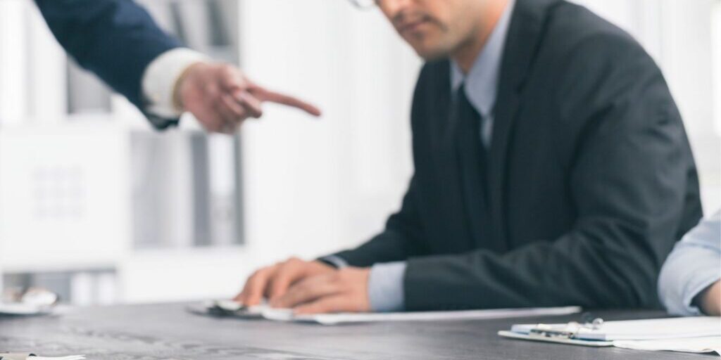 A blurry image of someone wearing a suit and glasses sitting at a meeting table and someone else on their right hand side pointing their index finger at them.