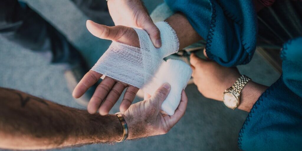 A photo of two people's hands. A person's right hand is being bandaged with a white bandage by another person to heal an injury.